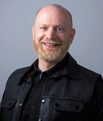 Picture of John smiling at the camera, wearing a black shirt and black leather sleeveless jacket